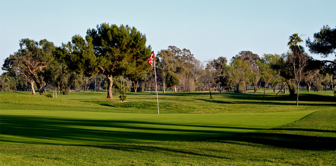 A golf course in Costa Mesa features a green with a flagstick marking the hole, trees in the background, and long shadows stretching across the grass under a clear sky—hinting at early morning or late afternoon.