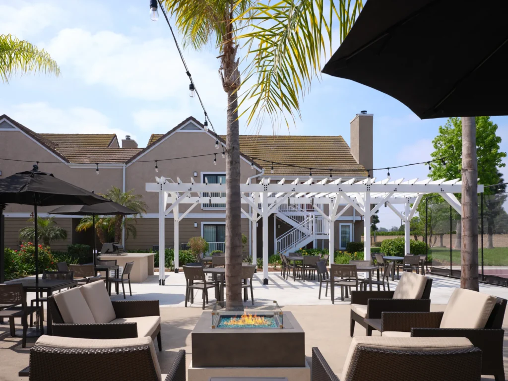 Outdoor patio with cushioned seating, tables, umbrellas, a fire pit, palm trees, greenery, and a white pergola in front of a tan building under a partly cloudy sky—an inviting setting that stands out among wedding venues Costa Mesa has to offer.