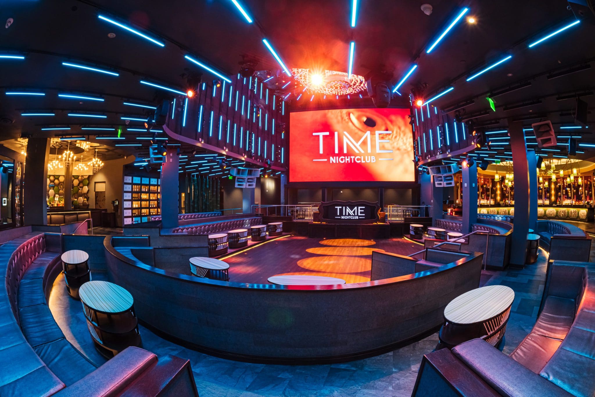 A modern nightclub interior in Costa Mesa with colorful LED lighting, a spacious dance floor, round booths, and a big TIME NIGHTCLUB screen above the stage—perfect for any Costa Mesa travel blog’s nightlife feature. The space is empty and well-lit.
