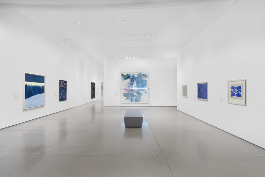 A modern art gallery with white walls displays colorful abstract paintings and photographs. A single gray bench is positioned in the center of the polished floor, facing a large blue artwork at the end of the room.