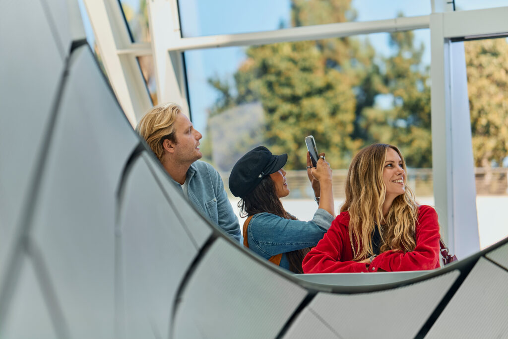 Three people stand indoors by large windows in a modern, bright setting; one takes a photo with a phone while the others smile and look around. Trees are visible outside, capturing a lively moment in news Costa Mesa CA style.