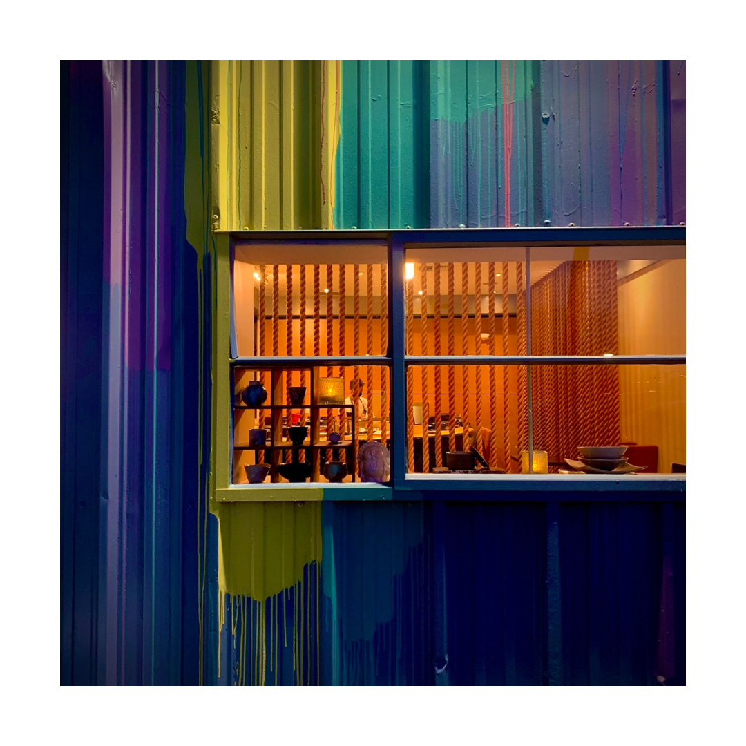 A colorful metal wall, painted with vertical streaks of yellow, green, blue, and purple, stands near Hana Re Sushi Costa Mesa, featuring a lit window that reveals shelves with pottery and warm light inside.