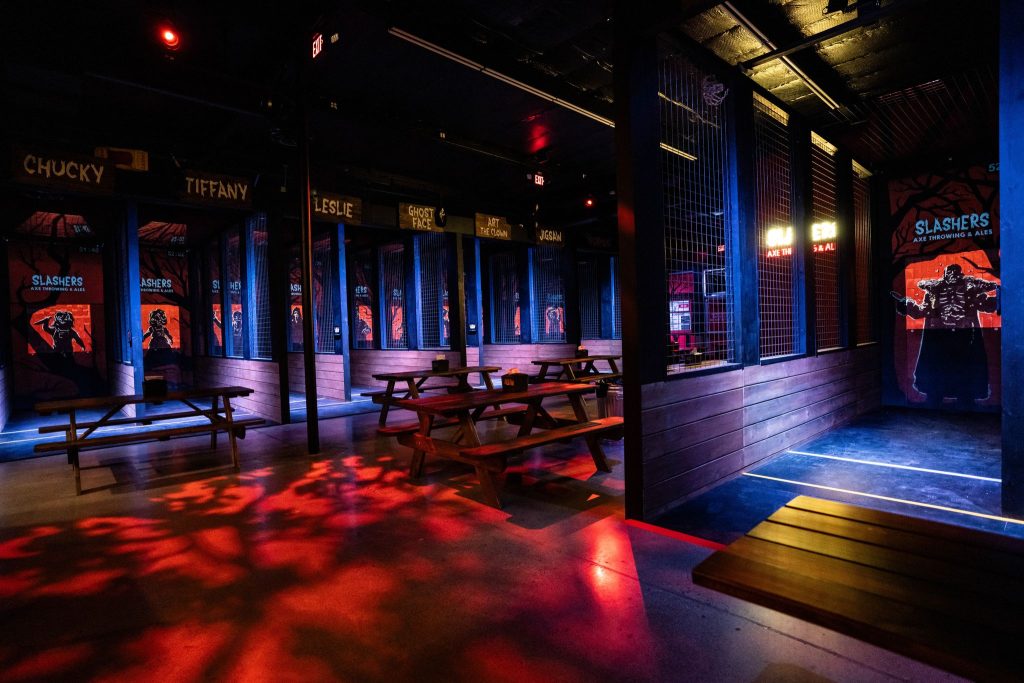 Dimly lit indoor venue with wooden picnic tables, dark walls, and axe-throwing lanes. Colorful red and blue lighting creates a dramatic atmosphere. Horror character posters and names are displayed above each lane.