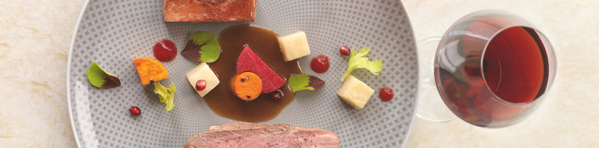 A gourmet dish featuring sliced meat, cubes of cheese, colorful vegetables, sauce, and garnish, artfully arranged on a patterned plate beside a glass of red wine at Knife Pleat Costa Mesa.