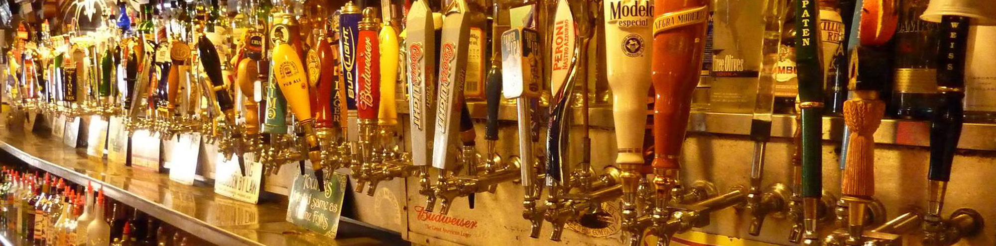 A long row of beer taps with colorful handles is lined up on the bar at Goat Hill Tavern Costa Mesa, with bottles and glasses visible below. The taps are closely arranged, offering a wide variety of draft beers.