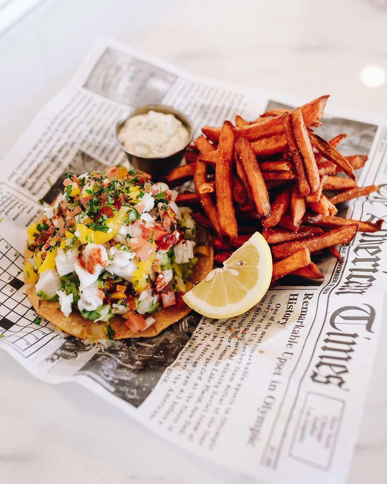 A plate from Tres Muchachos Costa Mesa: sweet potato fries, a lemon wedge, dipping sauce, and a colorful tostada with diced vegetables and herbs, all served on newspaper-style parchment.