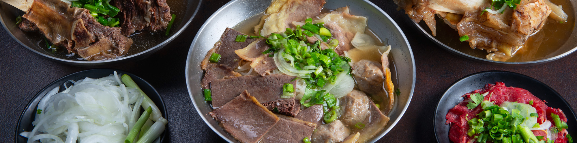 Five bowls arranged on a table at Phoholic Costa Mesa, featuring Vietnamese beef noodle soup with various cuts of beef, bone marrow, fresh herbs, sliced onions, and a dish of raw beef slices.