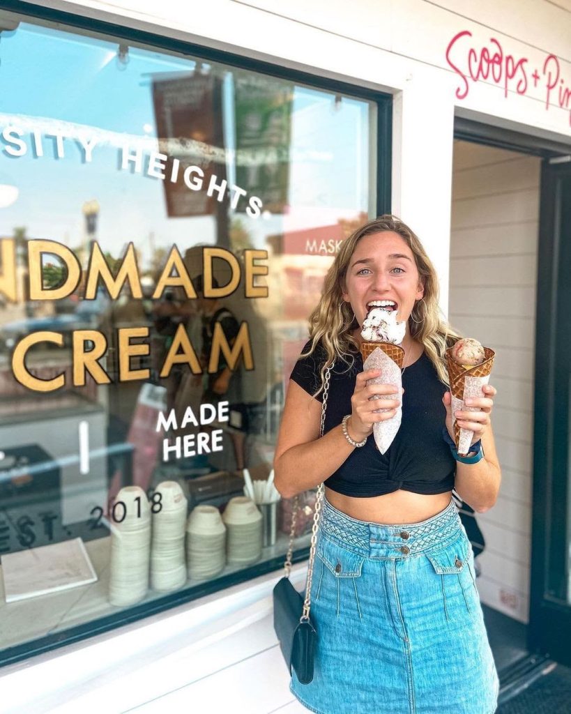 A woman in a black crop top and denim skirt stands outside an ice cream shop, smiling and holding two ice cream cones, one in each hand. The shop window behind her displays signs for handmade ice cream.