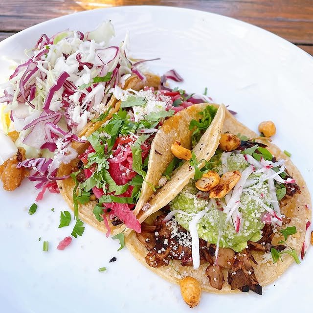 Two tacos topped with guacamole, pickled onions, cheese, roasted chickpeas, and fresh herbs are served on a white plate with a side of crispy fried pieces and colorful shredded cabbage slaw.