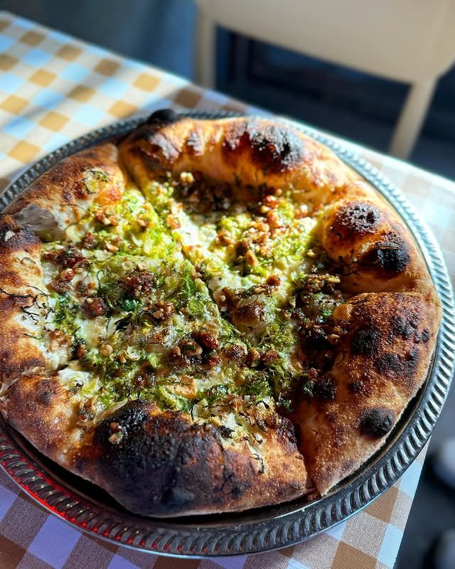 A close-up of a wood-fired pizza with a bubbly, slightly charred crust, topped with herbs, cheese, and a drizzle of olive oil, served on a metal plate on a checkered tablecloth.