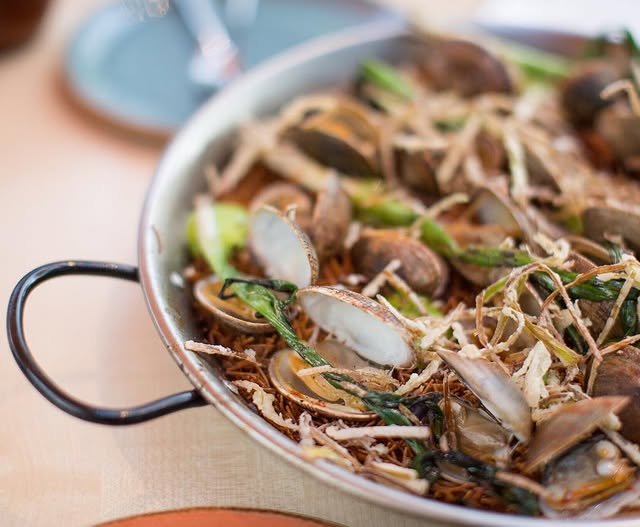 A close-up of a pan filled with clams, noodles, leafy greens, and crispy fried strips, served on a light wooden table with plates in the background.