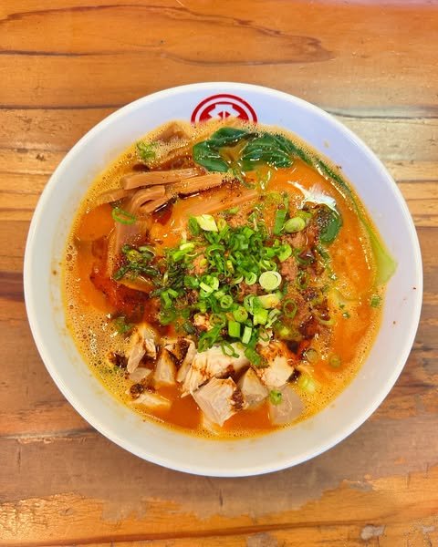 A bowl of spicy ramen topped with chopped green onions, sliced pork, bamboo shoots, and vegetables, set on a wooden table. The broth appears rich and orange-red with visible oil and seasonings.