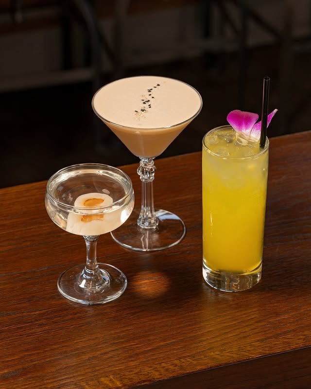Three cocktails on a wooden table: a clear drink with an olive in a coupe glass, a creamy martini with black garnish, and a tall yellow cocktail with ice, a straw, and a purple flower.