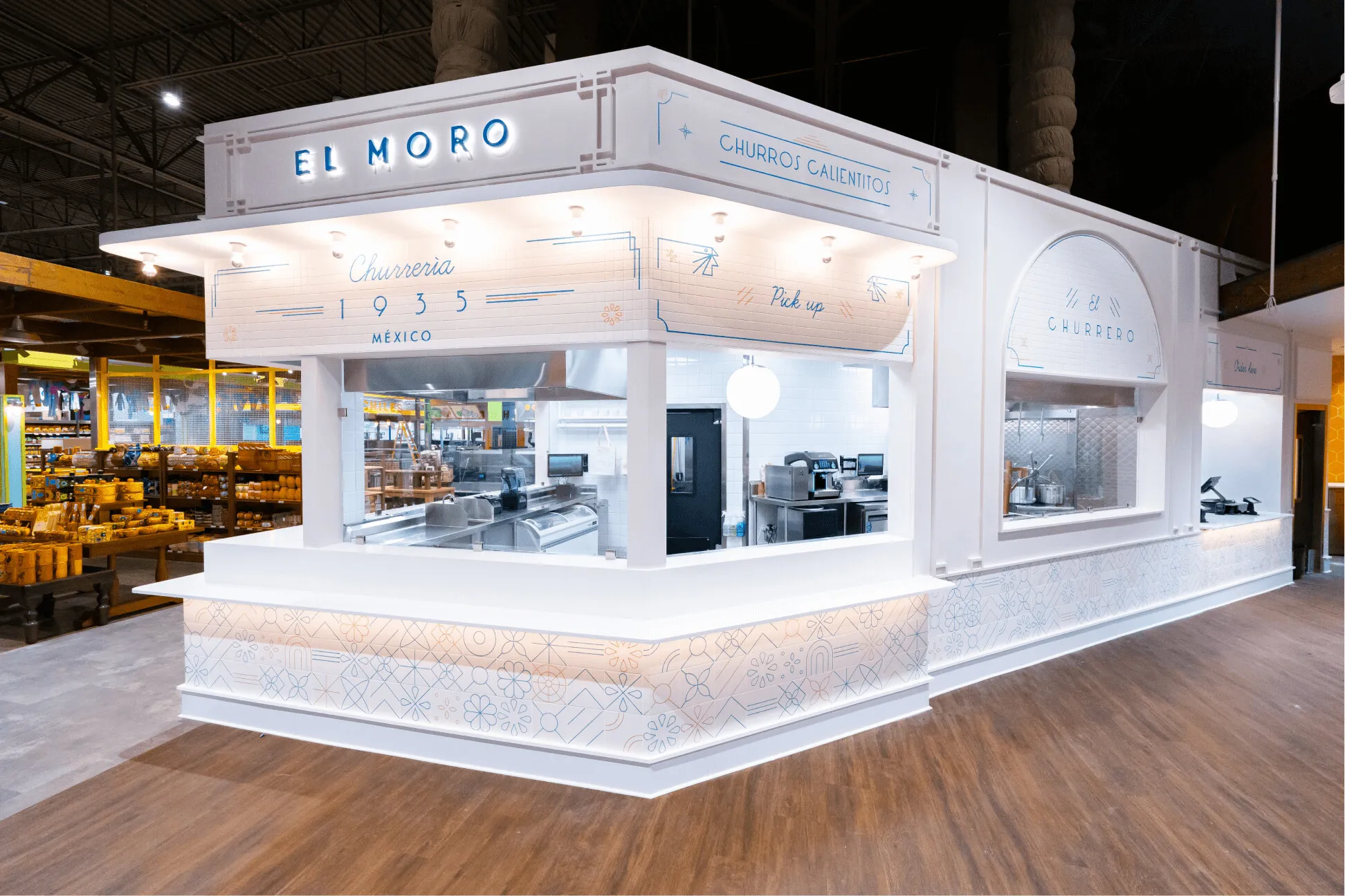 A modern, white churro stand named El Moro with light blue accents and a neon sign, located inside a spacious market. The counter is clean, and the kitchen area is visible behind the pick-up window.