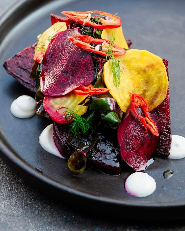 A colorful gourmet dish featuring roasted beetroot, thinly sliced yellow and purple root vegetables, red chili, fresh herbs, and dollops of white sauce, artfully arranged on a dark plate.