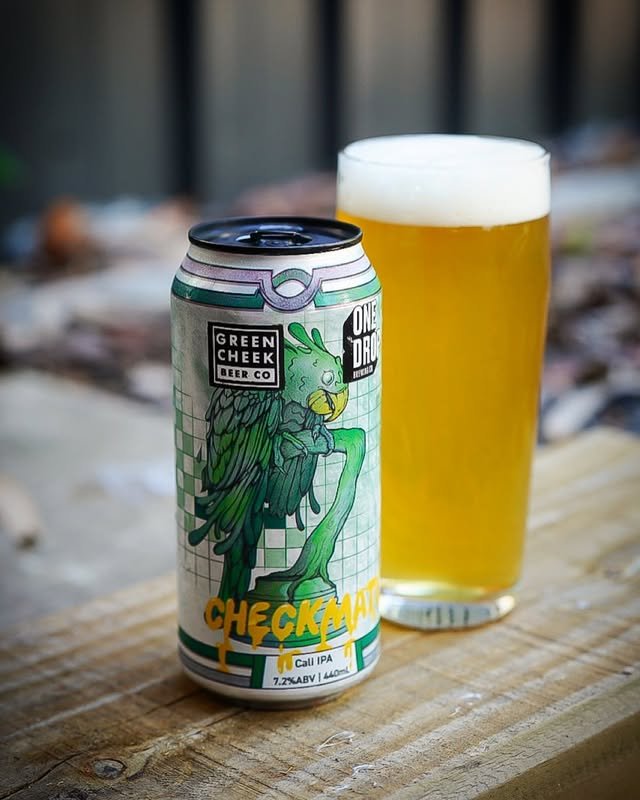 A can of Green Cheek Beer Co. and One Drop Brewing’s “Checkmate” Cali IPA sits next to a glass of hazy golden beer on a wooden surface, with a blurred outdoor background.