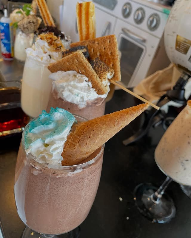 Three extravagant milkshakes topped with treats: one with whipped cream and a wafer cone, another with whipped cream, toasted marshmallow, and graham crackers, and a third with whipped cream and a churro, all on a black counter.