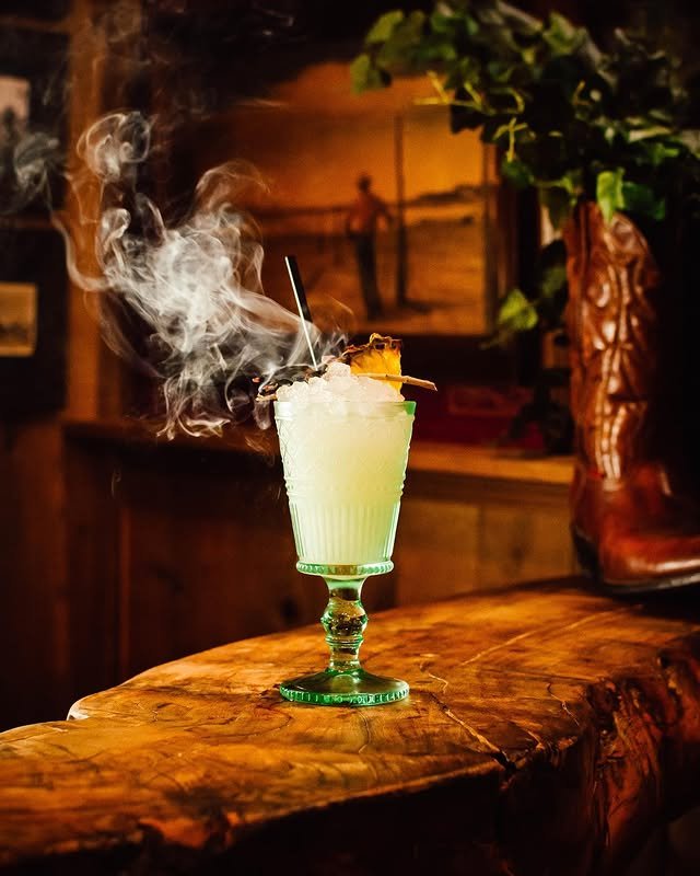 A green glass filled with a yellow cocktail, topped with crushed ice, garnish, and smoke rising from the drink, sits on a rustic wooden bar with warm lighting and blurred decor in the background.