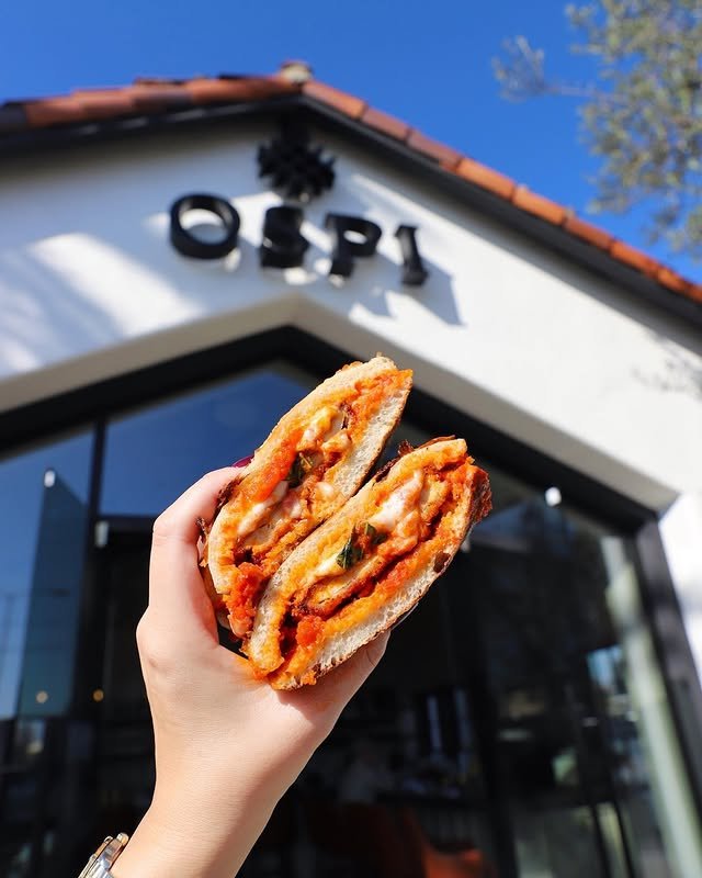 A hand holds up a sandwich filled with melted cheese, tomato sauce, and herbs in front of a building with the sign “OSPI” on it. The sky is clear and blue above the building.