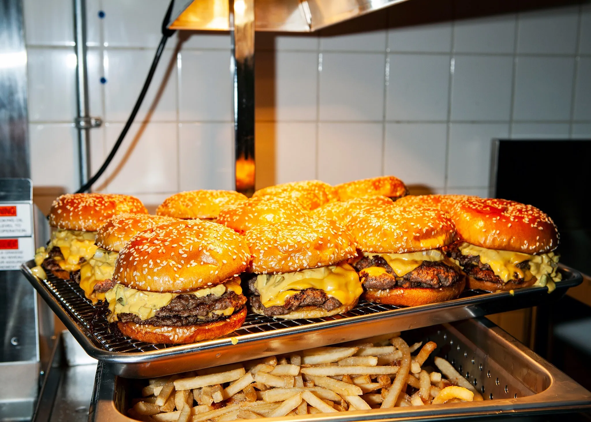 A tray of cheeseburgers with sesame seed buns is displayed above a container filled with French fries in a kitchen setting.