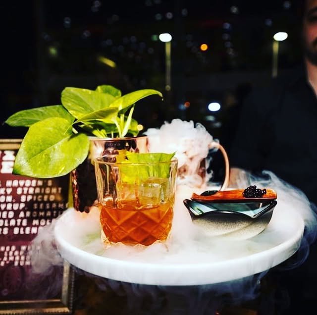 A fancy cocktail served on a round tray with dry ice mist, featuring a copper mug with leaves, a glass with an amber drink and mint, and a small dish holding a dessert garnished with berries.