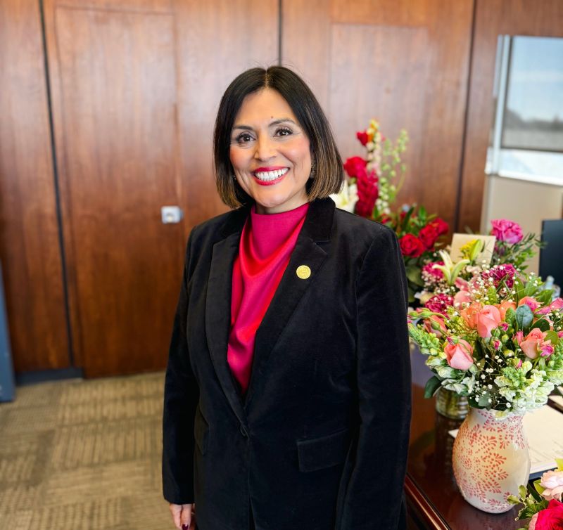A woman with shoulder-length dark hair, wearing a black blazer and red blouse, stands smiling in an office with wooden walls. A desk nearby holds vases of colorful flowers.