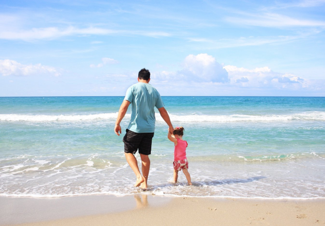 A man and a young child holding hands and walking along the shore of a sunny beach, with gentle waves and a blue sky in the background—moments like these await when you visit Costa Mesa.
