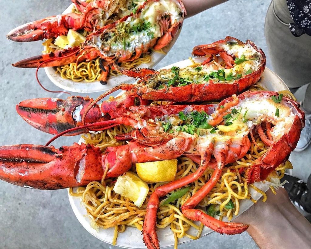 Two plates topped with grilled lobster halves on a bed of stir-fried noodles, garnished with chopped herbs and lemon wedges, held by people over a concrete surface.