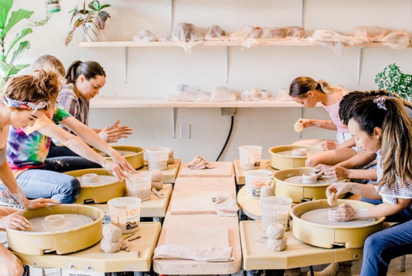 Five women sit at pottery wheels in a bright studio, shaping clay with their hands. Shelves with pottery and supplies are on the wall behind them, and plants are visible in the background.