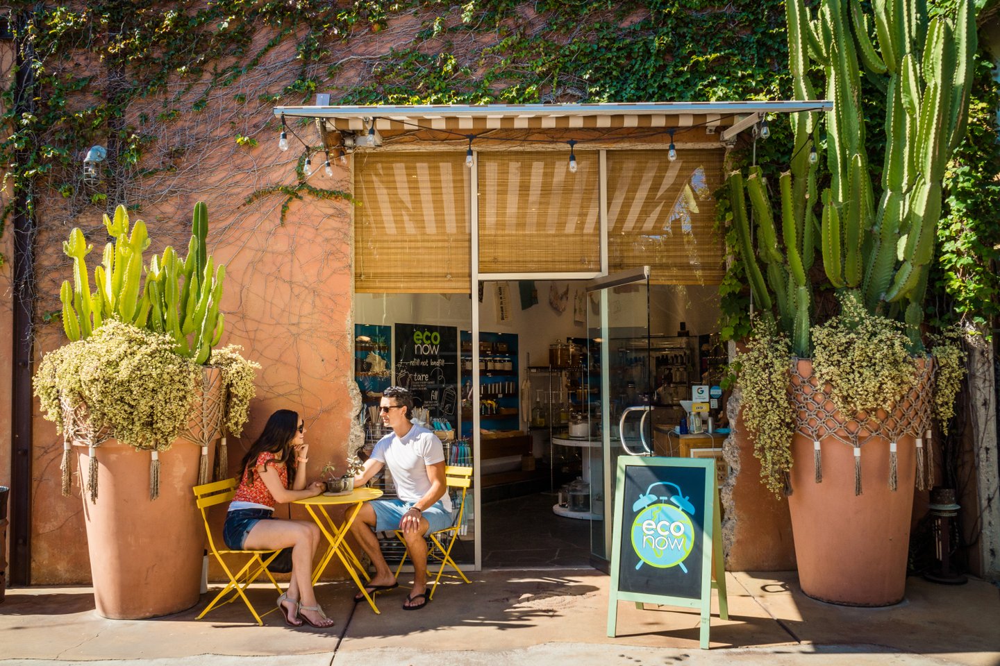 Two people sit at a small yellow table outside a shop called Eco Now, which is decorated with large potted cacti and leafy plants, under a striped awning on a sunny day.