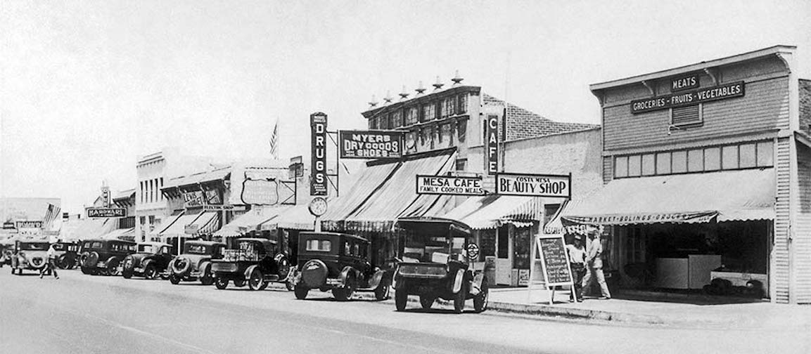 Black and white photo of a 1920s or 1930s downtown street with vintage cars parked by shops—like a cafe, drug store, and grocery market. People stroll the sidewalk, capturing the early history of Costa Mesa.