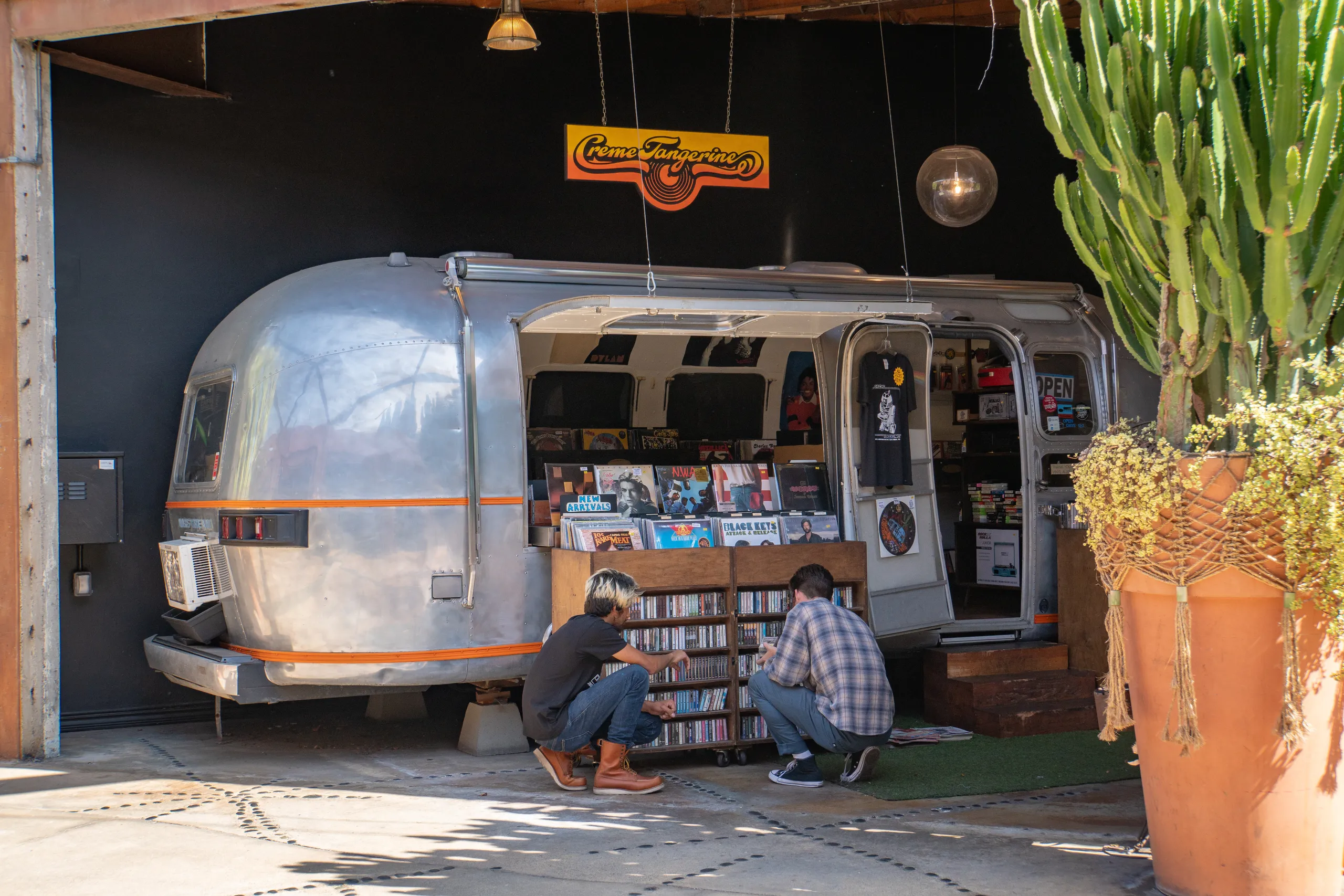 Two people crouch in front of a vintage silver trailer converted into a record store, browsing through vinyl records displayed on shelves. A large potted cactus stands nearby, and the store has a retro sign above it.