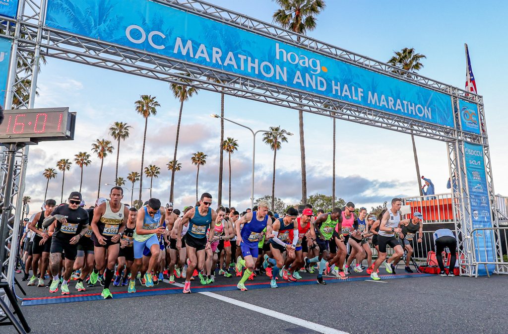 Runners begin the race at the starting line of the OC Marathon and Half Marathon, with palm trees and a blue sky in the background, under a large banner displaying the event name.