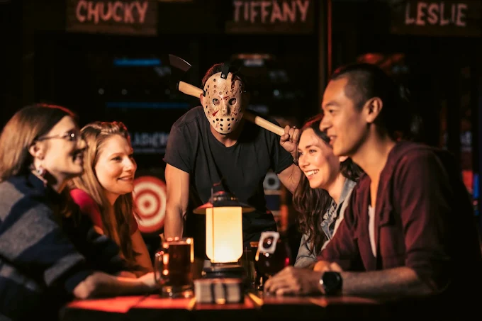 A group of four people sit at a table laughing, while a person behind them wears a hockey mask and holds an axe over their shoulder; the scene is warmly lit and playful.