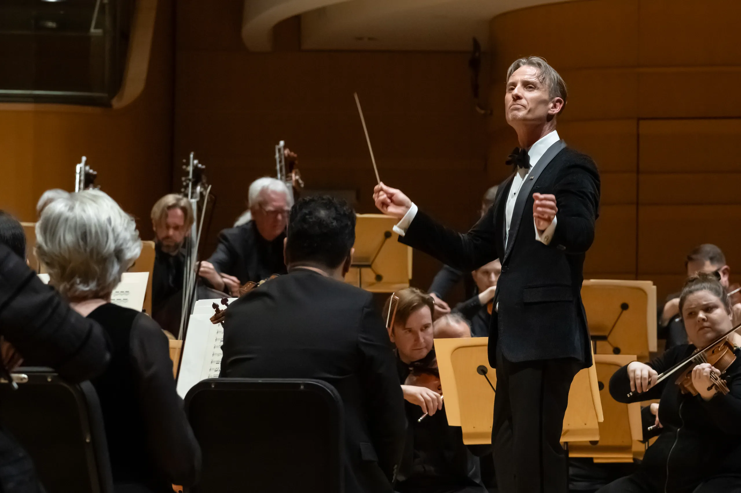 A conductor stands on stage in front of the Pacific Symphony Orange County, holding a baton and leading musicians who are seated and playing string instruments in a concert hall.