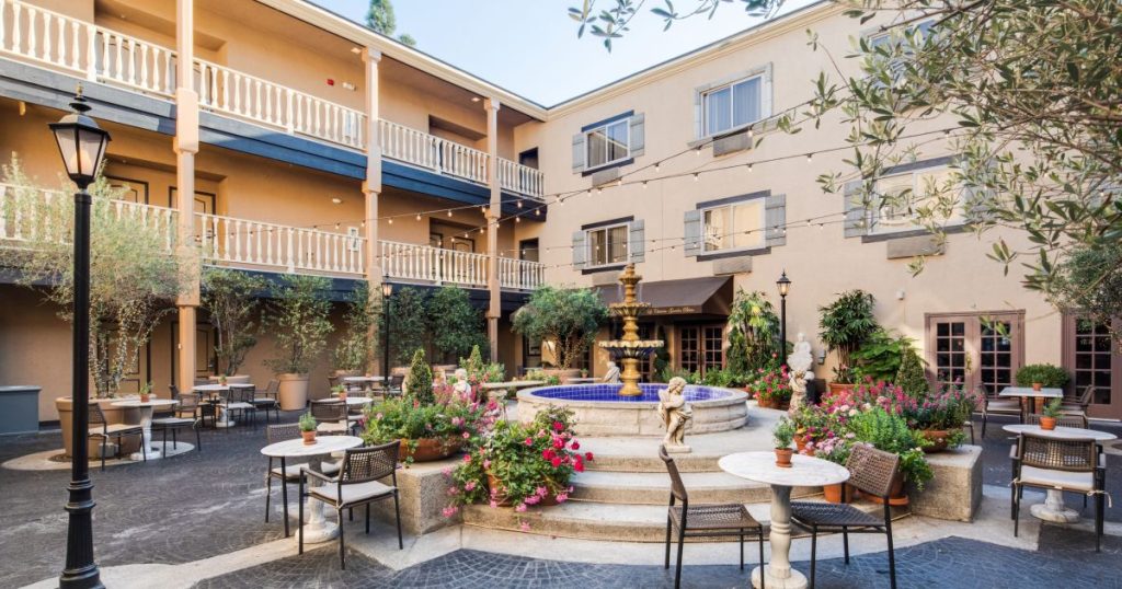 Outdoor courtyard with round tables and chairs, a central tiered fountain surrounded by flowers and greenery, and a three-story building with balconies and large windows in the background.