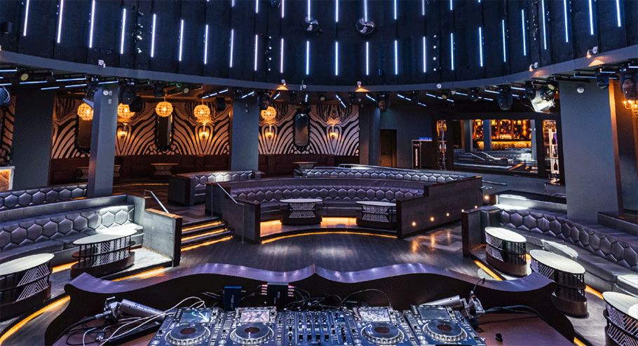 A modern nightclub interior with a DJ booth in the foreground, curved seating areas, round tables, geometric wallpaper, and blue accent lighting—an ideal choice among meeting venues Costa Mesa has to offer.