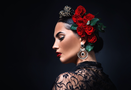 A woman in profile with dramatic makeup, wearing a black lace top, large ornate earring, jeweled headpiece, and red roses in her dark hair against a dark background.