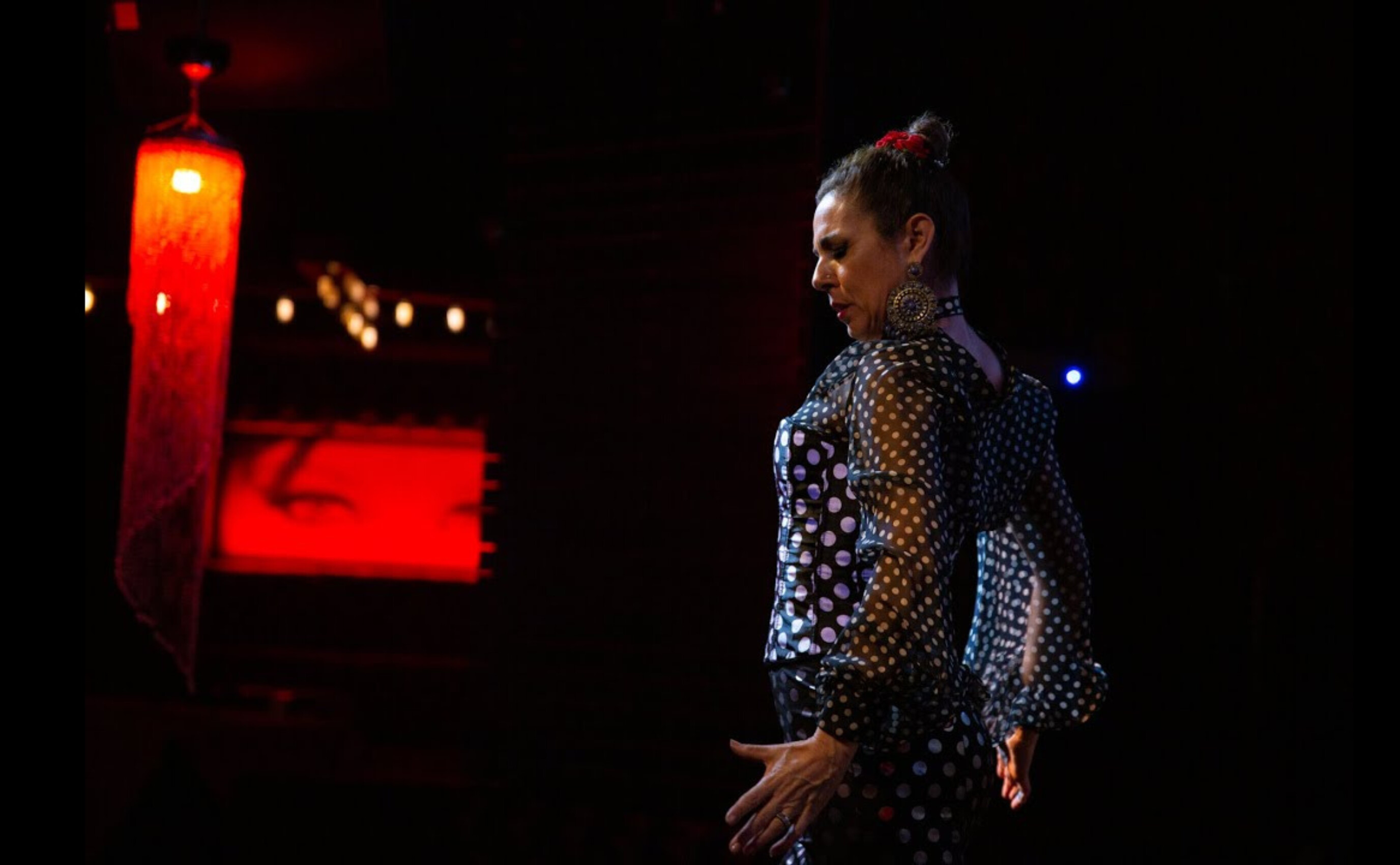 A flamenco dancer in a black dress with white polka dots performs under dim lighting, with a red lantern hanging above and a red-lit background featuring a pair of eyes.