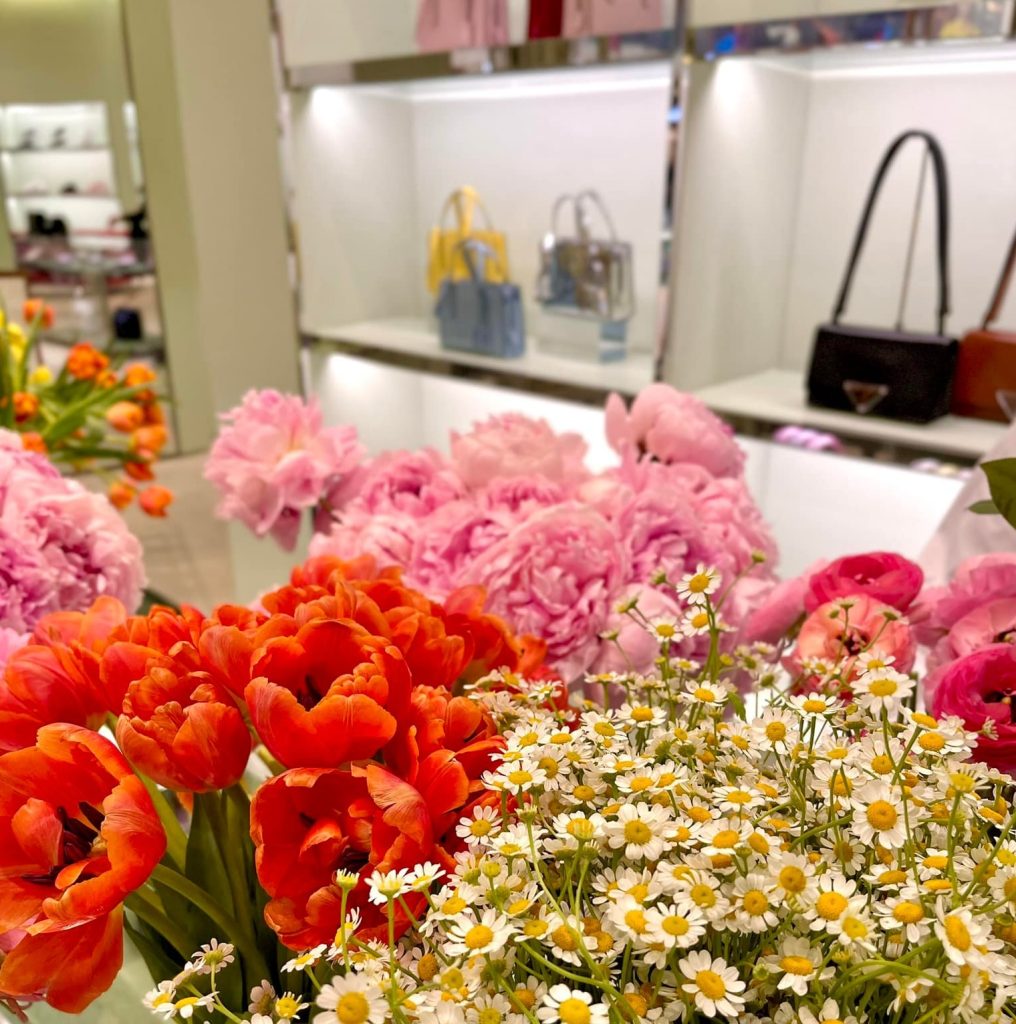 A display of vibrant flowers, including orange tulips, pink peonies, and white daisies, is set in front of shelves with designer handbags in a well-lit boutique.