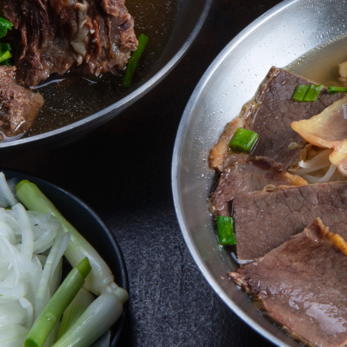 Close-up of a bowl of beef noodle soup with slices of beef, green onions, and broth. Next to it is a bowl of fresh white noodles—just the kind you might find at Michelin star restaurants in Orange County—set on a dark surface.
