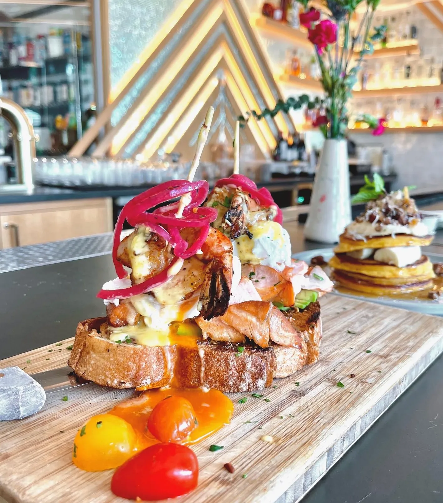 Open-faced sandwich on toasted bread topped with roasted vegetables, meat, pickled onions, and sauce, garnished with cherry tomatoes on a wooden board—perfect for your next Costa Mesa travel blog feature in a modern, stylish restaurant setting.