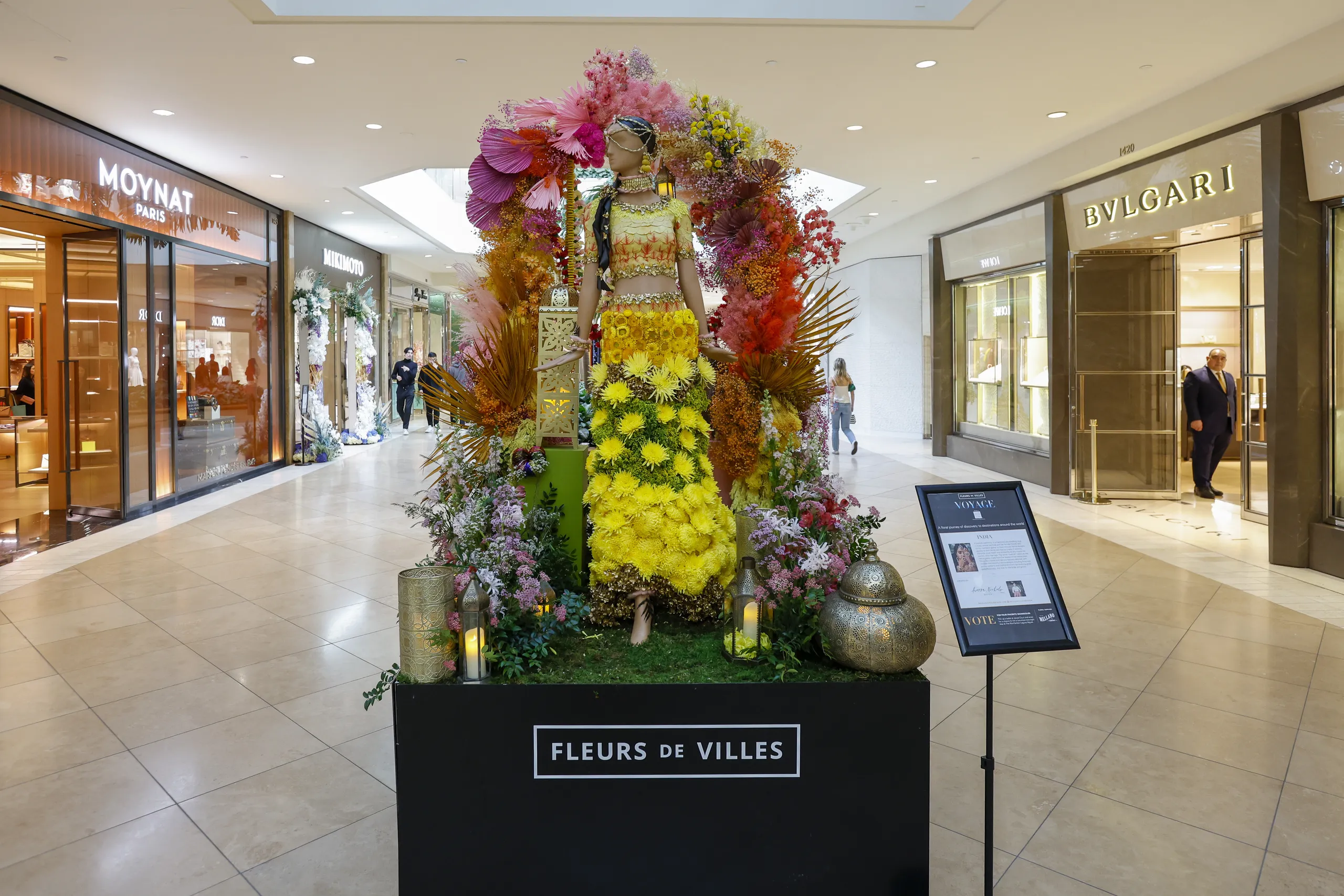 A mannequin decorated with vibrant flowers and foliage stands on a display labeled “Fleurs de Villes” in a shopping mall, surrounded by luxury store fronts and shoppers in the background.