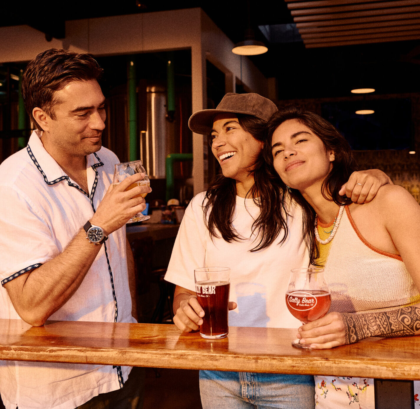 Three friends stand at a bar in Costa Mesa, smiling and holding drinks. One person leans on another, and they all appear relaxed and happy in the warmly lit indoor setting.