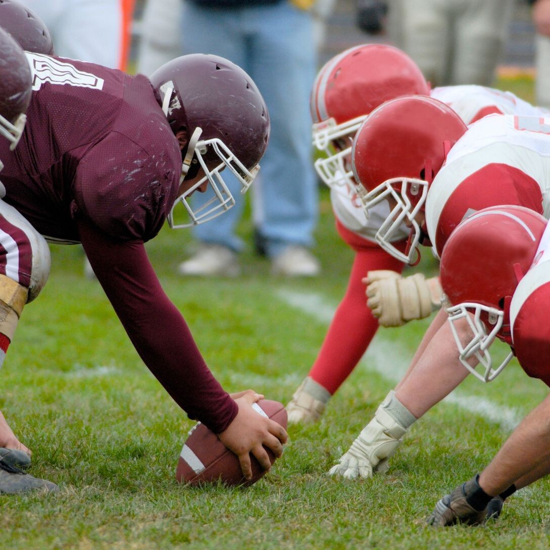 Football players from two teams crouch at the line of scrimmage, ready for a play. This intense moment unfolds on a field that rivals top event venues in Costa Mesa, as a maroon-uniformed player prepares to snap the ball against opponents in white and red.