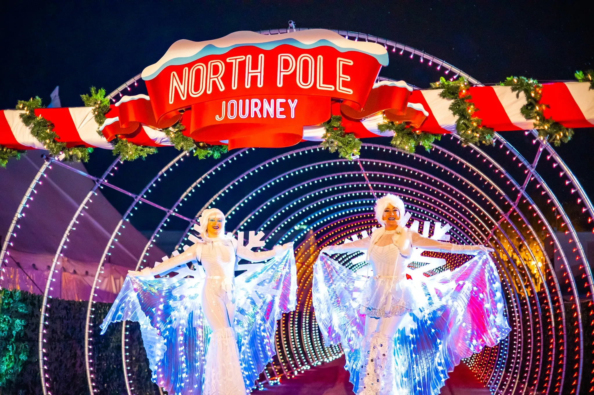 Two performers in glowing, festive costumes stand under a bright red and white “North Pole Journey” sign, surrounded by illuminated arches and holiday decorations at night—a magical moment perfect for any Costa Mesa travel blog.