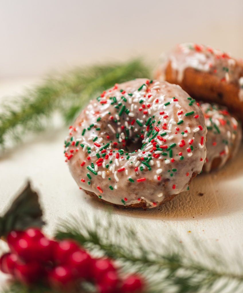 A glazed donut topped with red, green, and white sprinkles sits on a white surface, surrounded by pine branches and red berries, creating a festive holiday atmosphere.