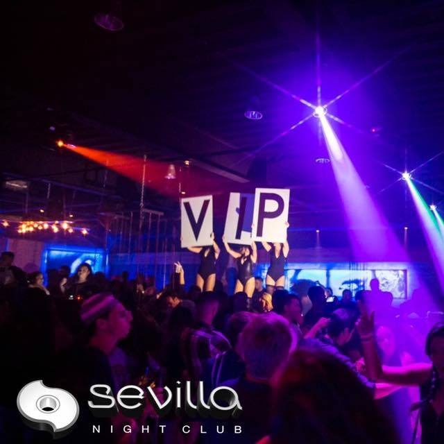 People dancing in a crowded nightclub with three performers on stage holding large signs that spell VIP. Colorful lights illuminate the scene. Sevilla Night Club logo is visible in the bottom left corner.