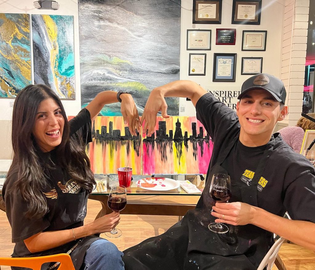 Two people sit side by side, smiling and holding glasses of red wine. Behind them is a colorful painting of a city skyline. They mimic the paintings shape with their arms, and art supplies are visible on the table in front of them.