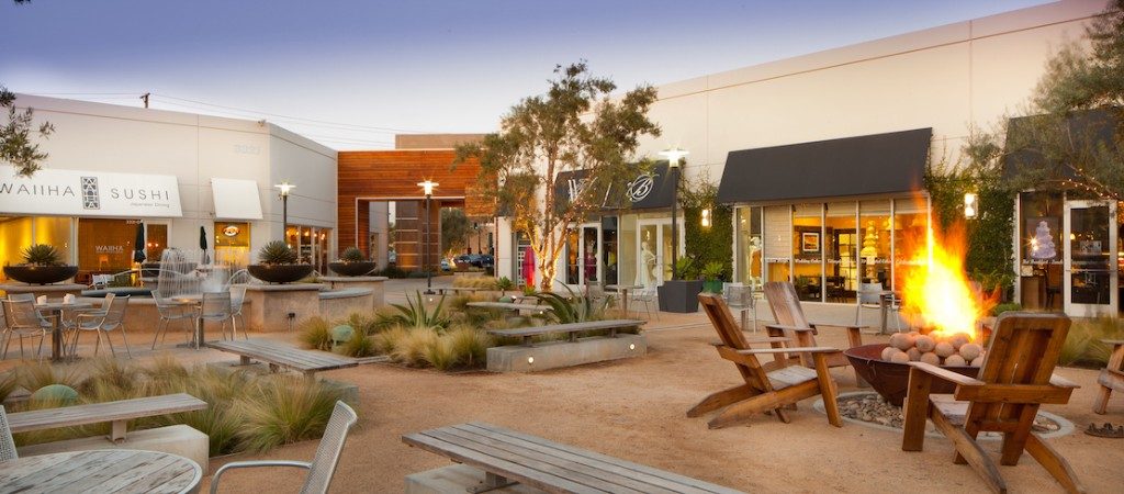 Outdoor shopping plaza with wooden benches, Adirondack chairs around a fire pit, plants, and small shops including a sushi restaurant. Warm evening light creates a welcoming vibe—ideal for gatherings or exploring wedding venues in Costa Mesa.